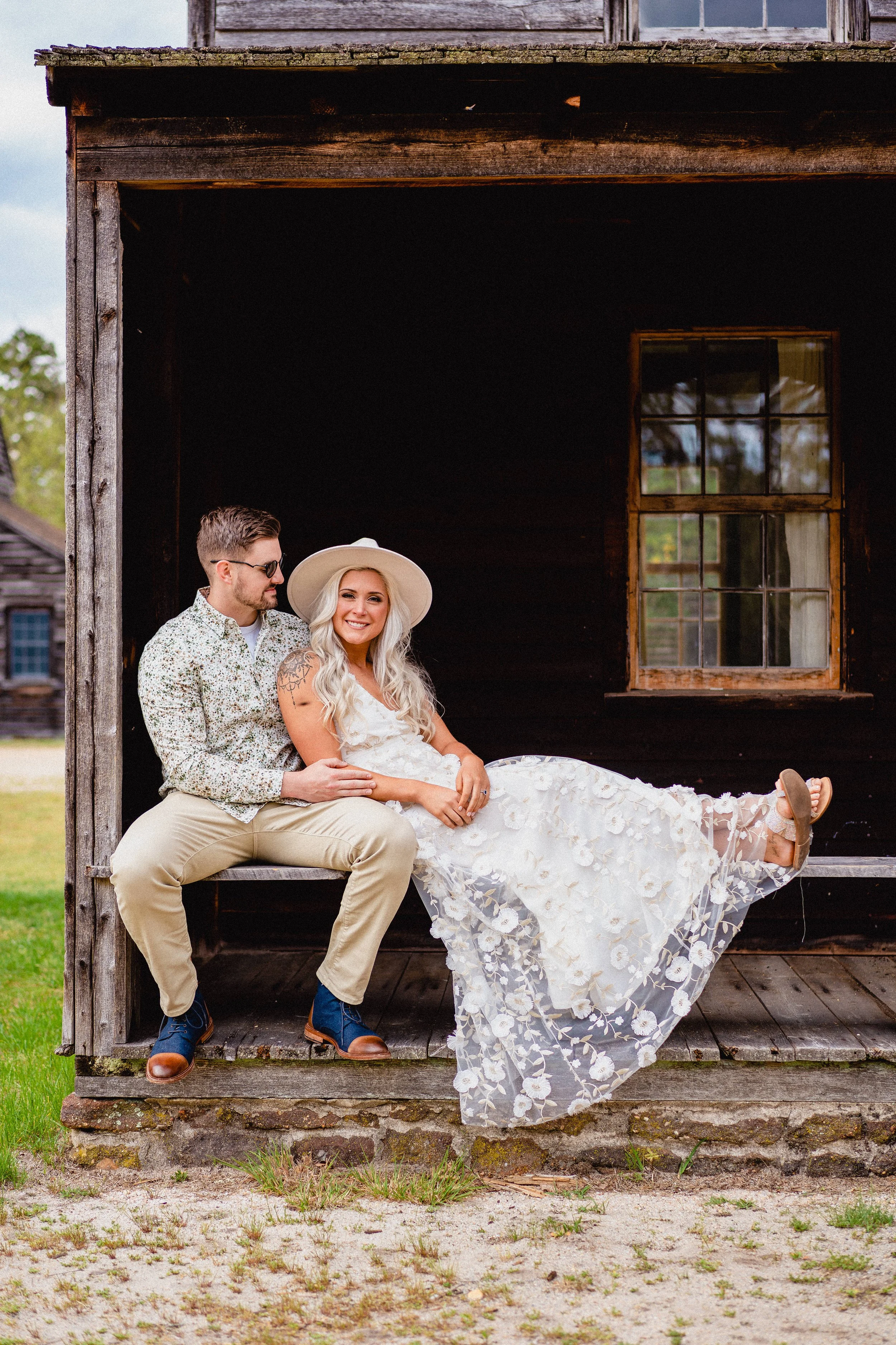 NJ Wedding Photography of A couple sitting on a wooden bench outside an old black barn. The woman is smiling, wearing a white dress with floral embroidery and a wide-brimmed hat. 