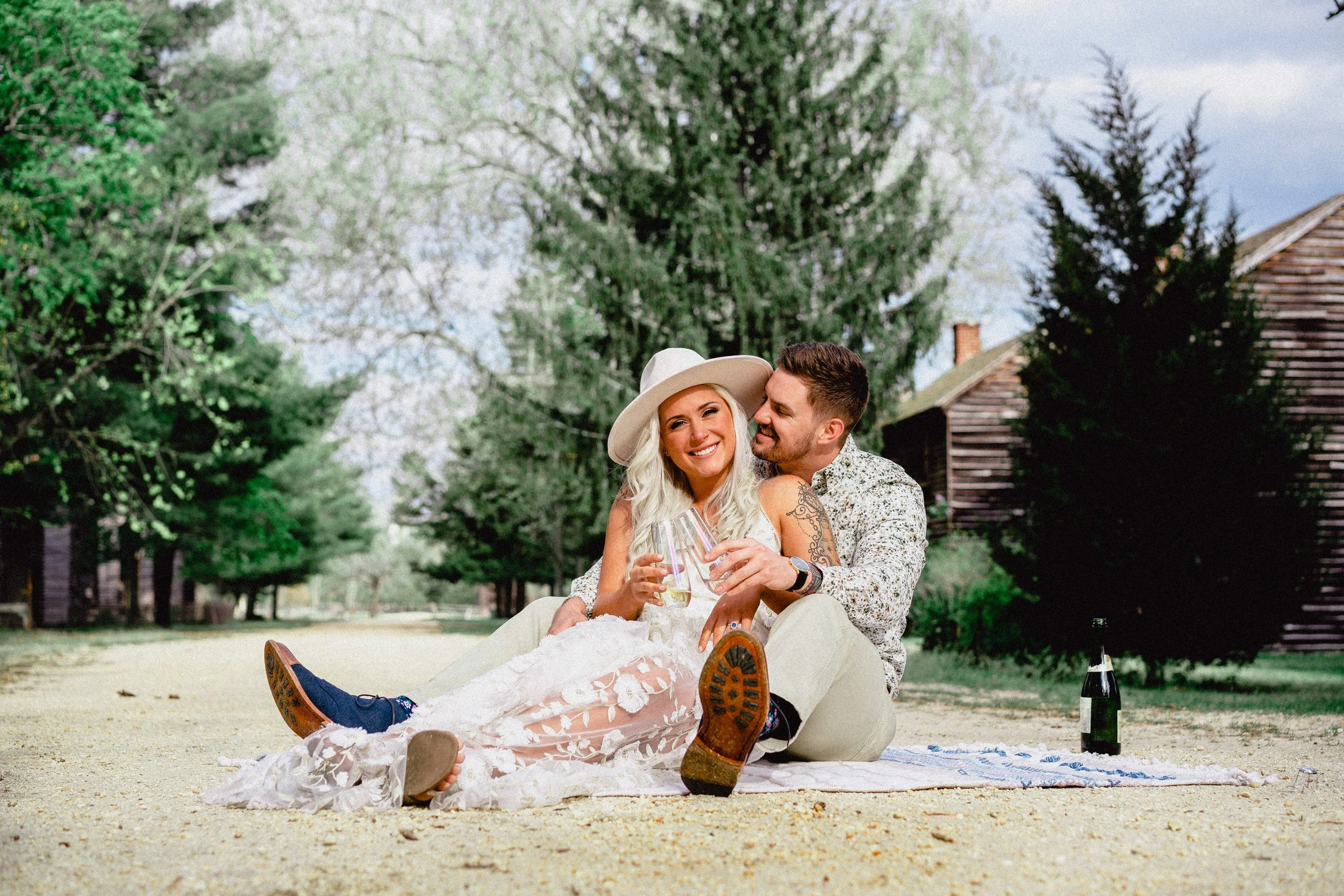 NJ Wedding Photography of A couple sitting on a blanket outdoors, holding glasses of wine, smiling and embracing, surrounded by trees and a rustic building, celebrating with a bottle of wine nearby.