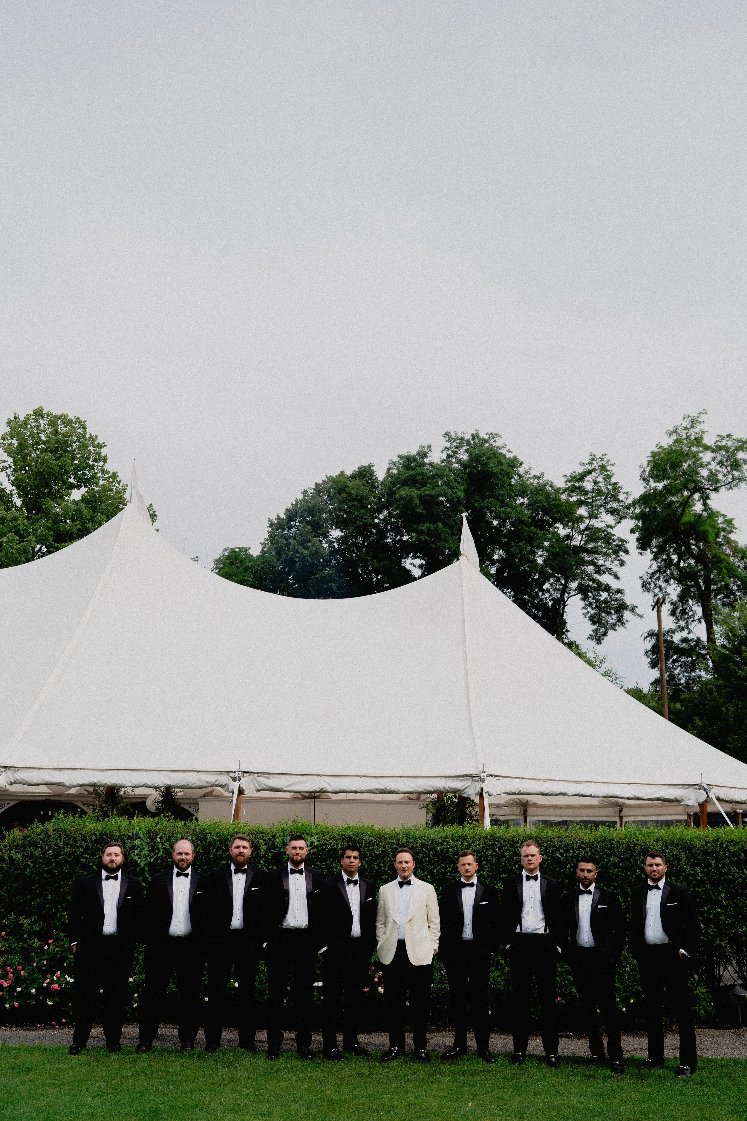 Black-and-white wedding photography of groomsmen lined up outside a luxury tent at an Upstate New York wedding.