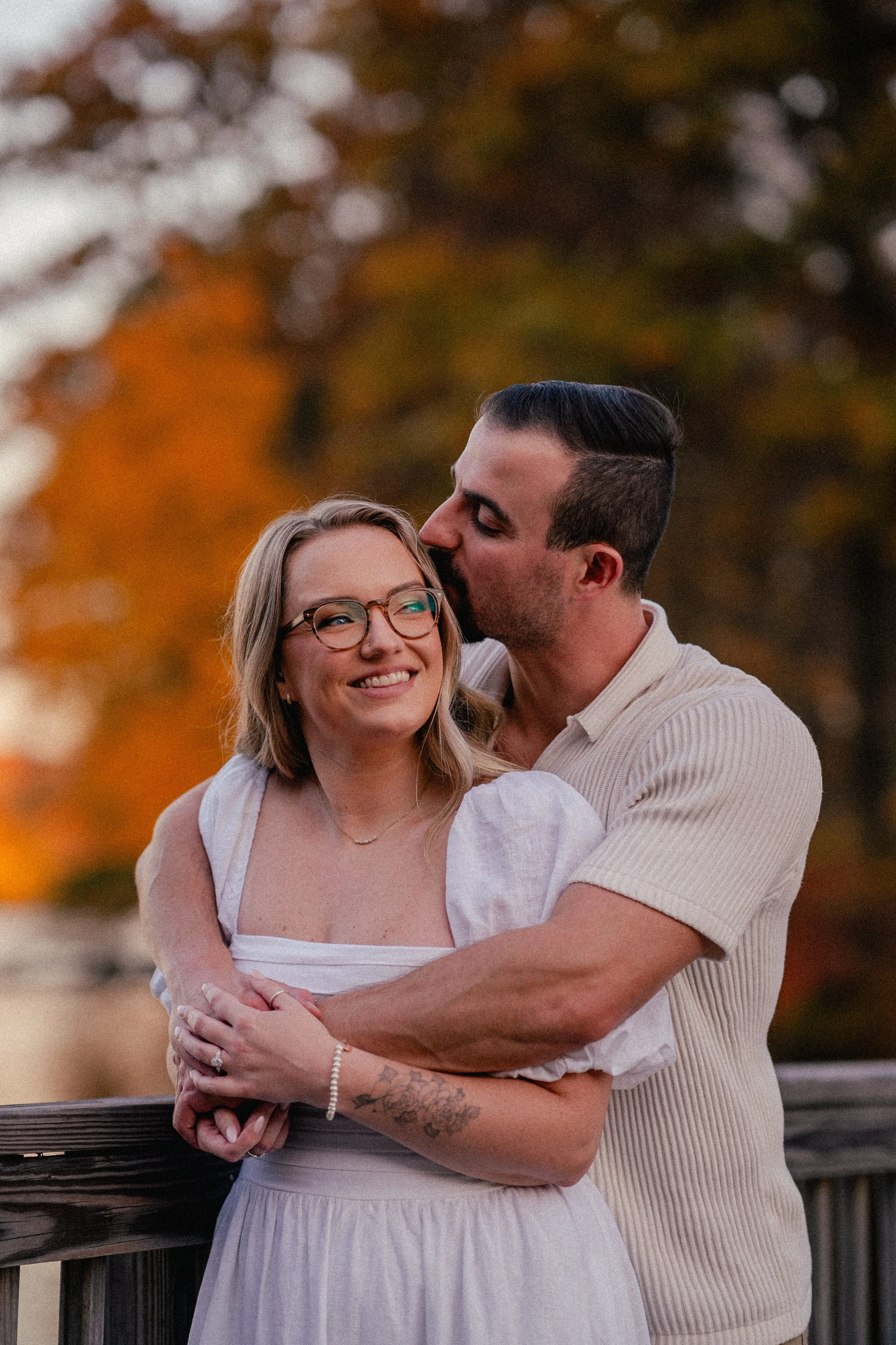 NJ Wedding Photography of A couple embracing on a wooden deck with autumn trees in the background. The woman is smiling, wearing glasses and a white dress, while the man kisses her on the head.