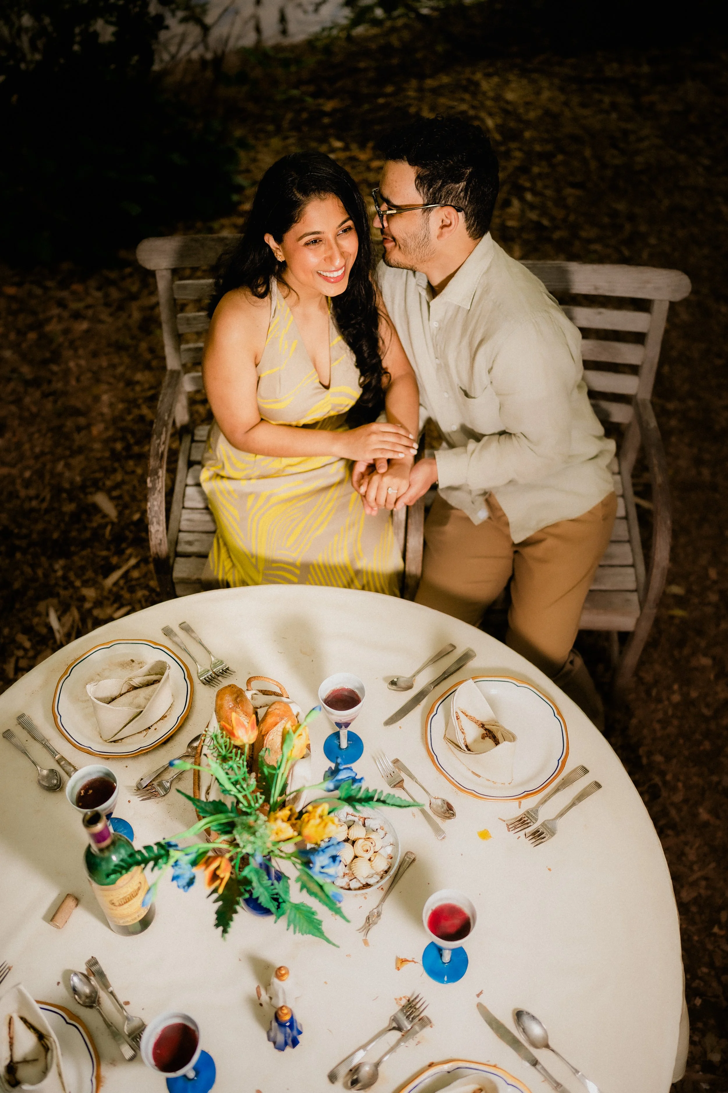 NJ Wedding Photography of A smiling woman and man holding hands and sitting close together on a bench at an outdoor dinner table at night.