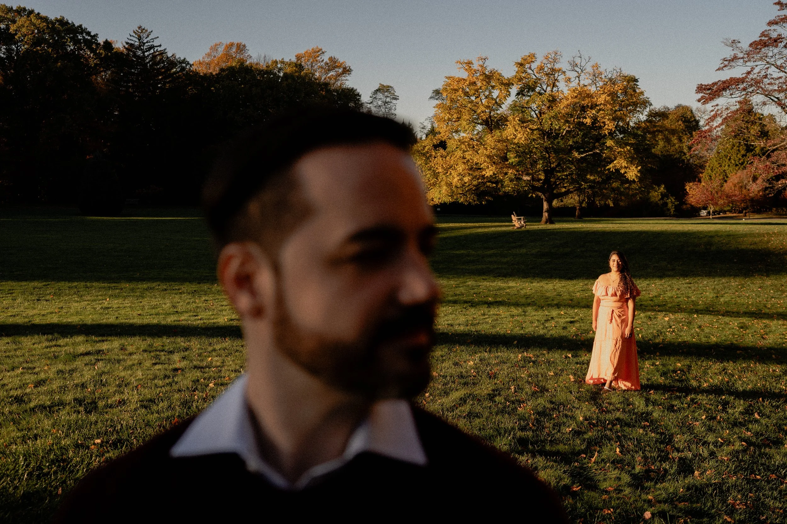 NJ Wedding Photography of A man in the foreground with a blurred face and a woman in the background standing in a park with trees and grass.