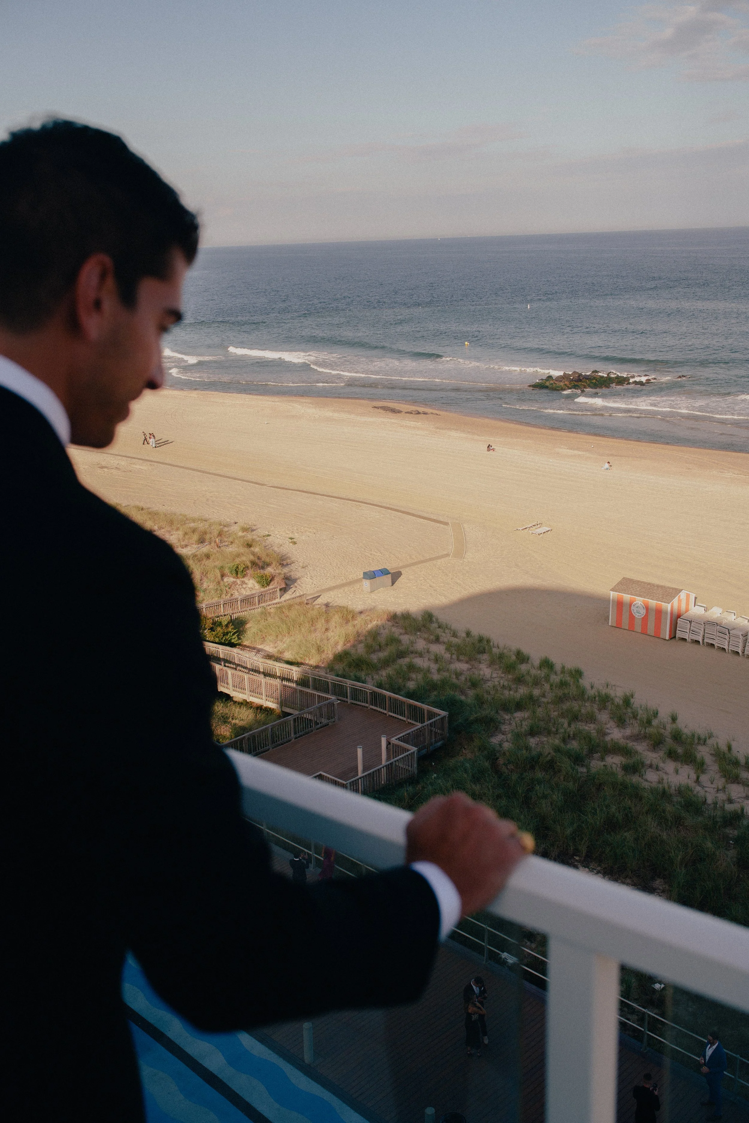 NJ groom standing on a balcony overlooking a beach with sand and the ocean. The man is facing sideways, looking down, with a partial view of his face and hand resting on the balcony railing at Wave resort - NJ Wedding photographers- Timel