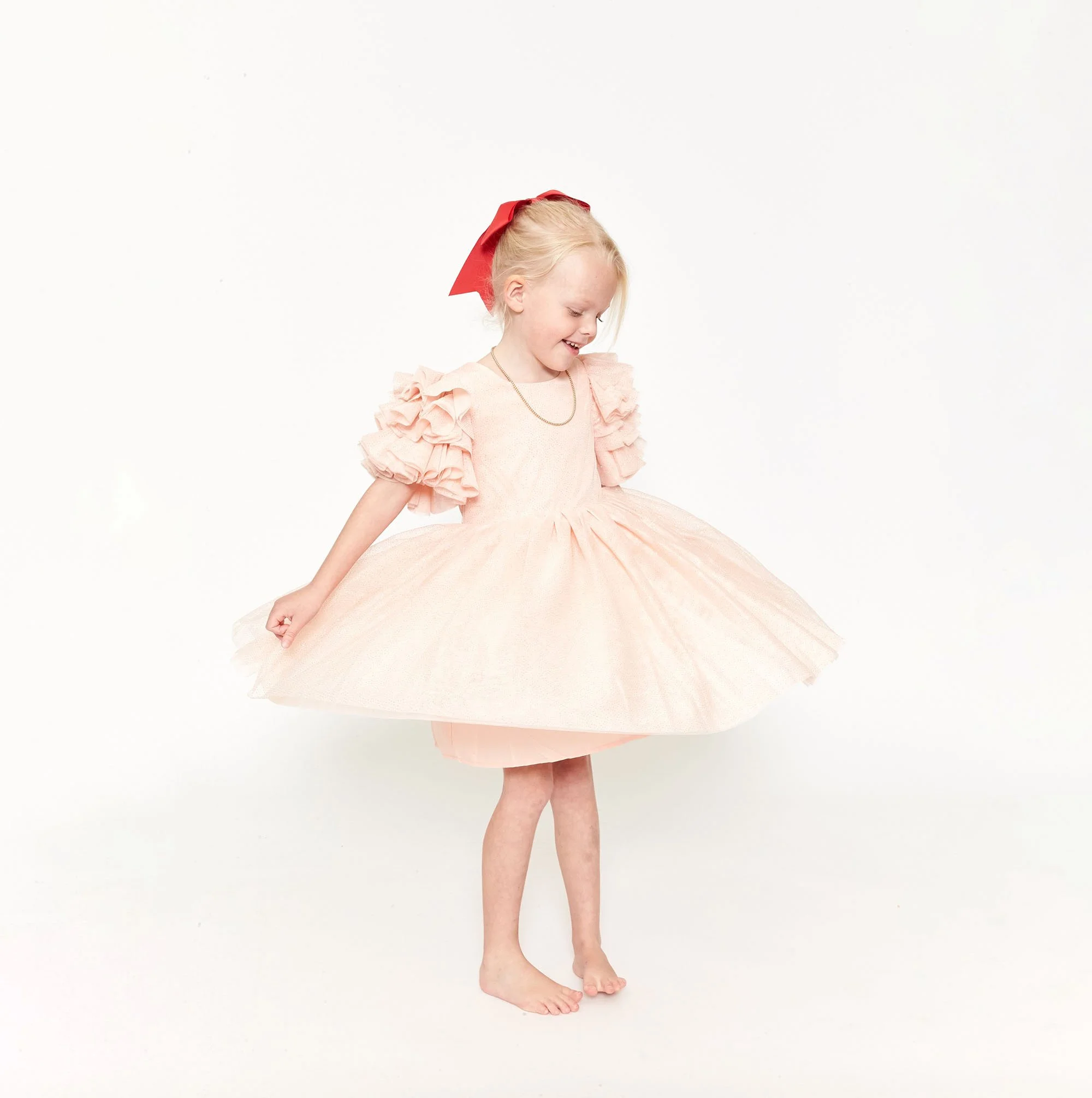 Young girl in a pink princess dress with puffed ruffled sleeves, a full skirt, and a red bow in her hair, smiling and looking downwards against a plain white background.