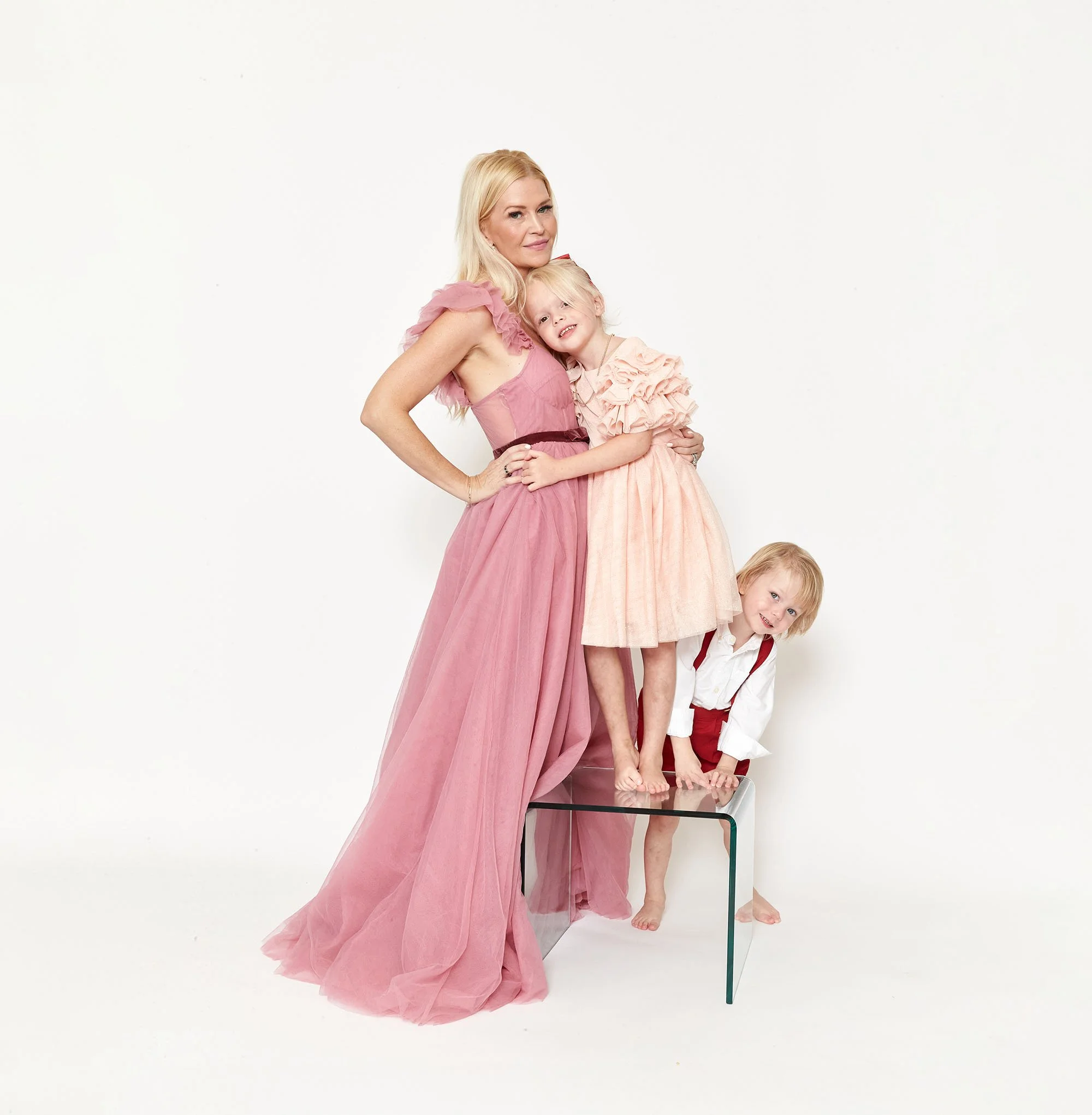 A woman and two children with blonde hair, dressed in pink and white, standing on and around a glass table in a white studio. The woman wears a pink dress, one girl wears a pink dress, and the boy wears a white shirt with red suspenders.