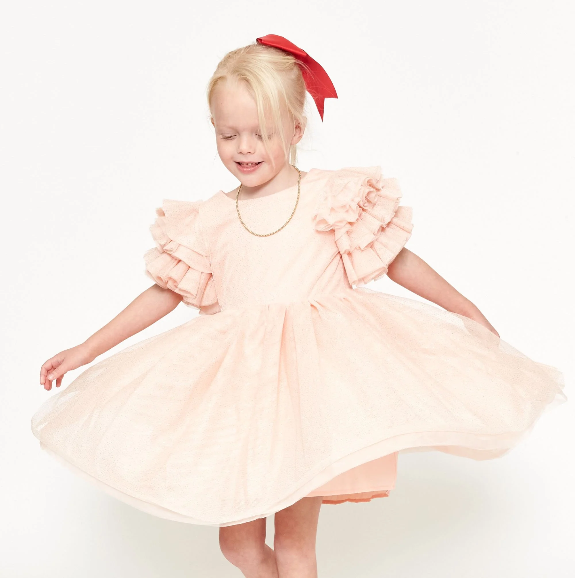 A young girl wearing a light pink dress with ruffled sleeves and a red bow in her blonde hair, holding out her skirt with a joyful expression.