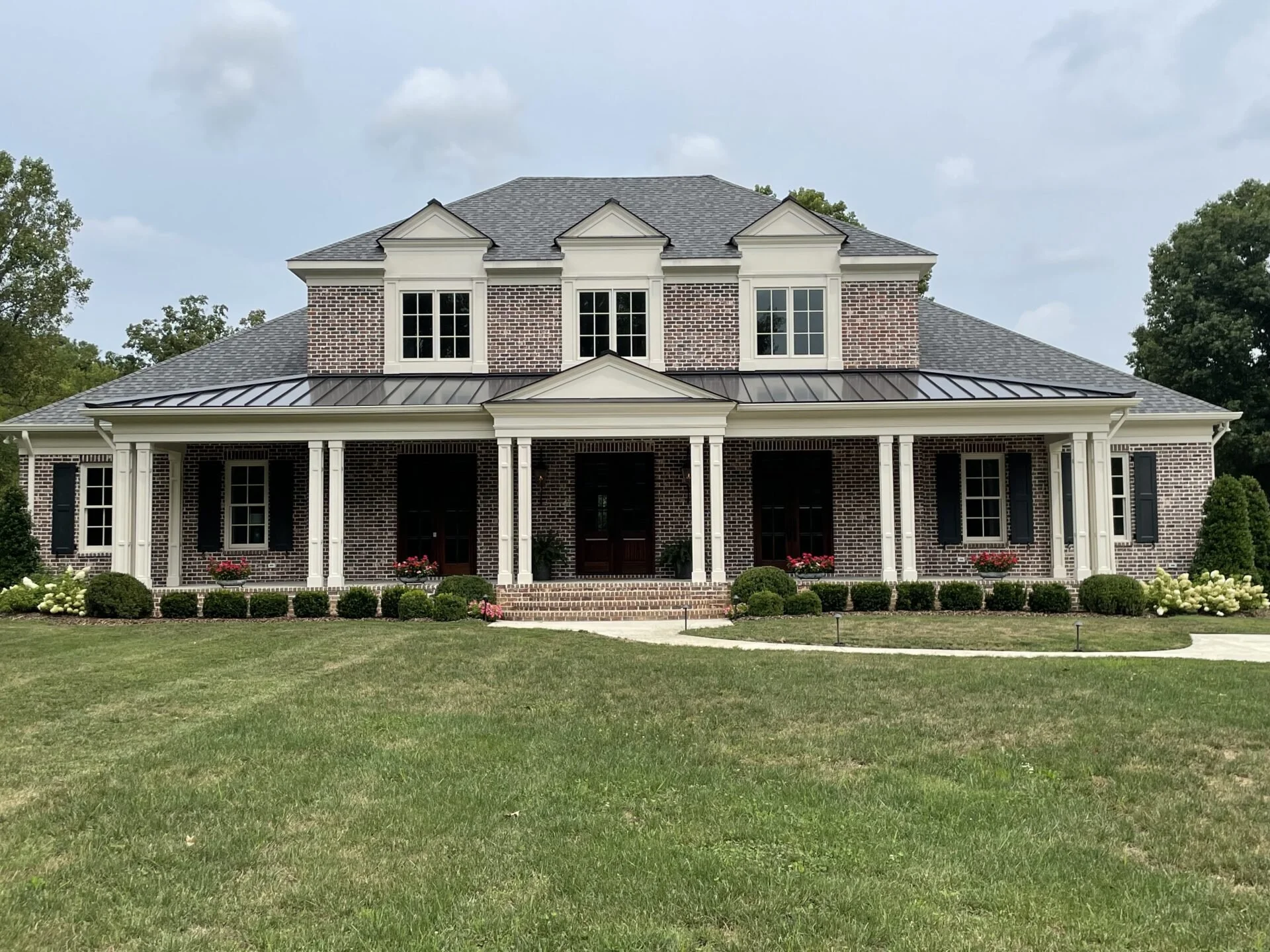 A large, two-story brick house with a front porch, white columns, and black shutters. The house has a gray roof, multiple windows, and landscaped front yard with bushes, flowers, and a walkway.