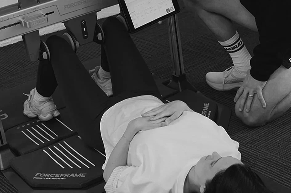 Person lying on a force frame testing device, with another person kneeling beside them, in a gym or lab setting.