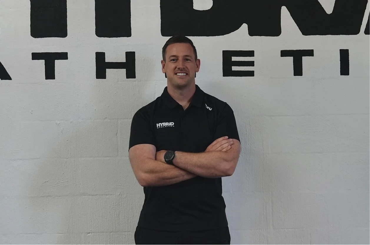 Man standing in front of a wall with black letters, arms crossed, wearing a black polo shirt with the words 'HYBRID ATHLETICS' on it.