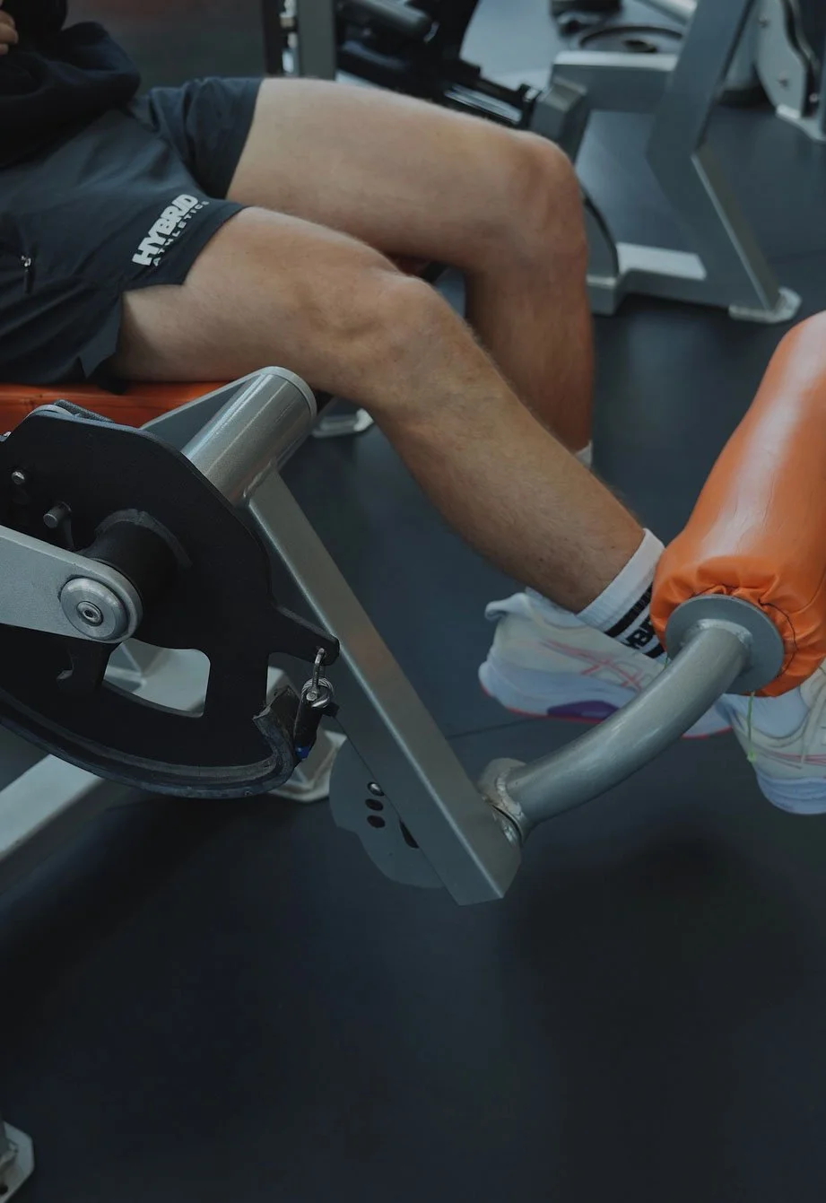 Person lying on a workout bench in a gym, wearing black athletic shorts and white sneakers, with their legs extended and feet secured under padded rollers.