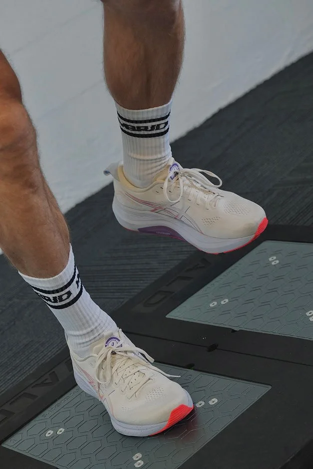 Close-up of a person's feet on a stair step in athletic shoes, white socks with black lettering, in a gym setting.