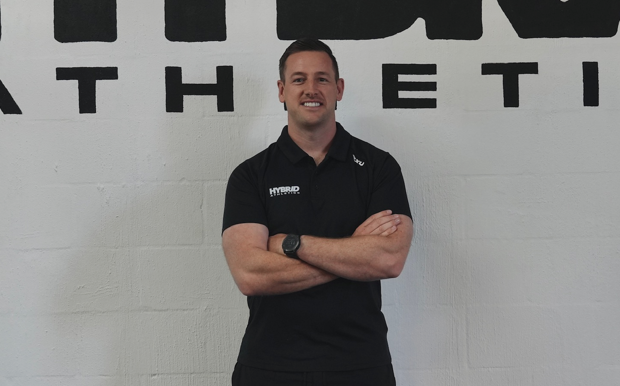 A man with short dark hair, wearing a black polo shirt with logos, stands with arms crossed in front of a wall with black letters, smiling at the camera.