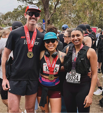 Three runners at a race event, wearing medals and race bibs, smiling in an outdoor setting with trees and other participants in the background.