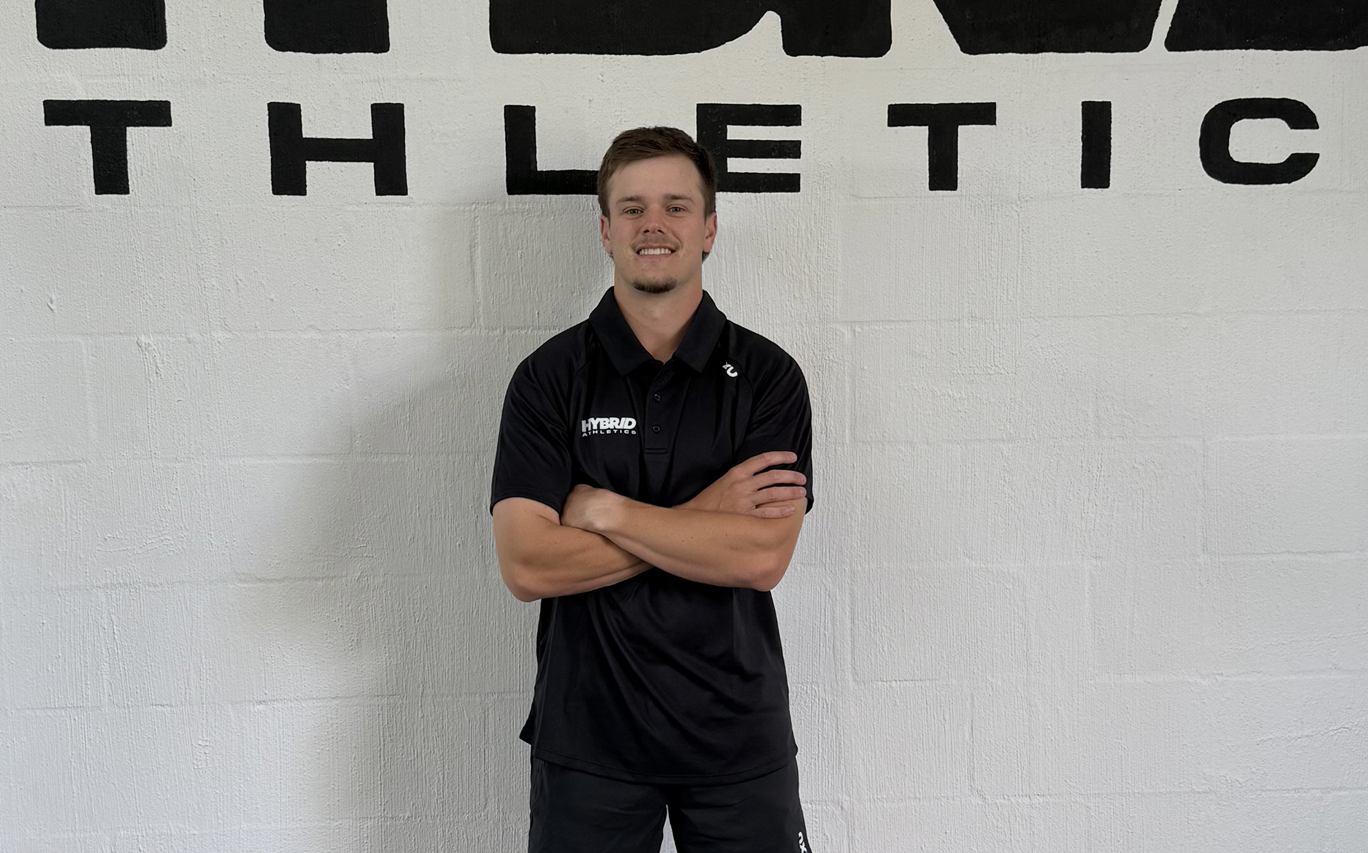 A young man standing with arms crossed in front of a white wall with black letters. He is wearing a black polo shirt with logos and text, and black pants, smiling at the camera.