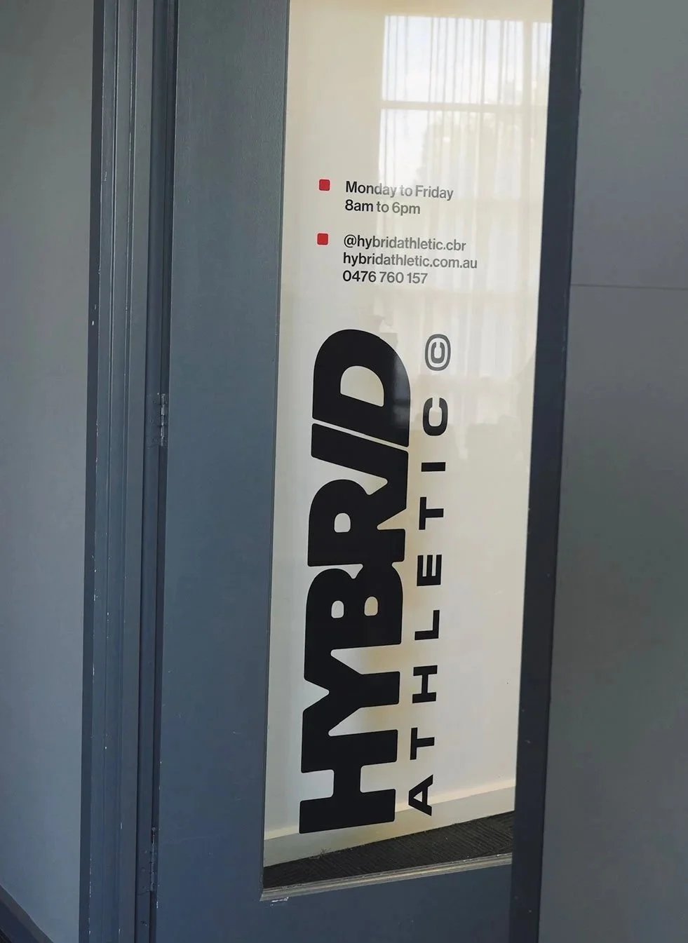 Glass door with a large black logo that reads 'HYBRID ATHLETIC' and a smaller sign with business hours, contact info, and social media handle.