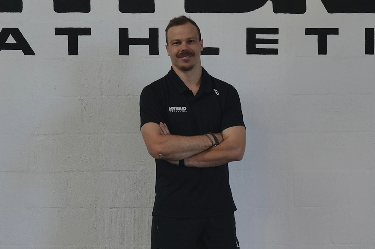 A man with a mustache and goatee standing with arms crossed in front of a white wall with black letters spelling "ATHLETIC." He is wearing a black polo shirt with a logo on the left side that says "HYBRID" and "ATHLETIC" and has several bracelets on his left wrist.