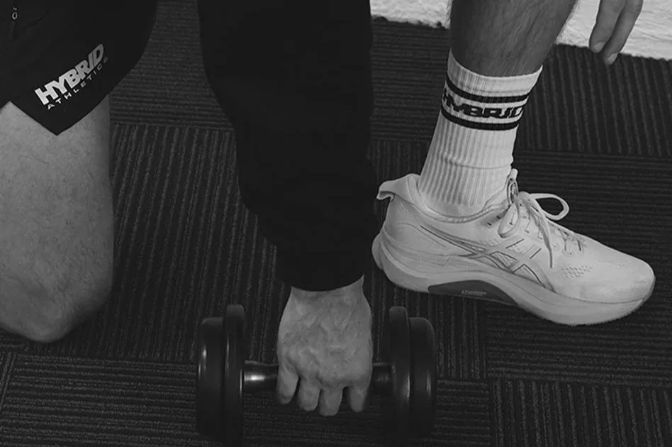 A person holding a dumbbell with their right hand, wearing a white athletic shoe, white sock, and dark pants, on a striped carpeted floor.