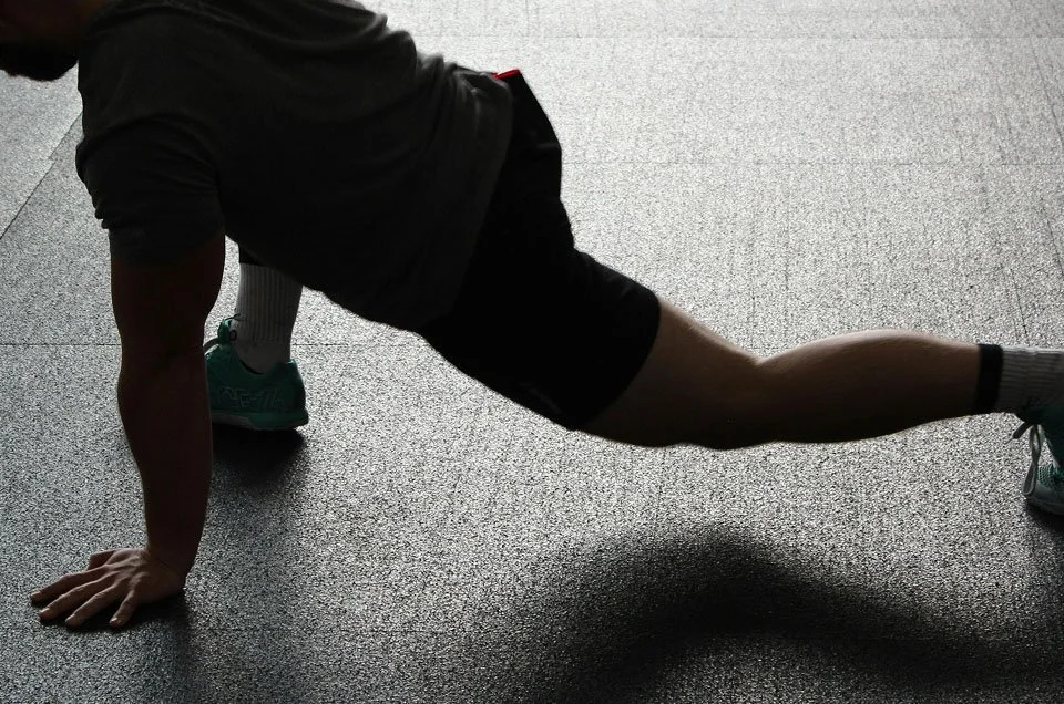 A person performing a stretching exercise on a textured gym floor, wearing athletic clothes and shoes.