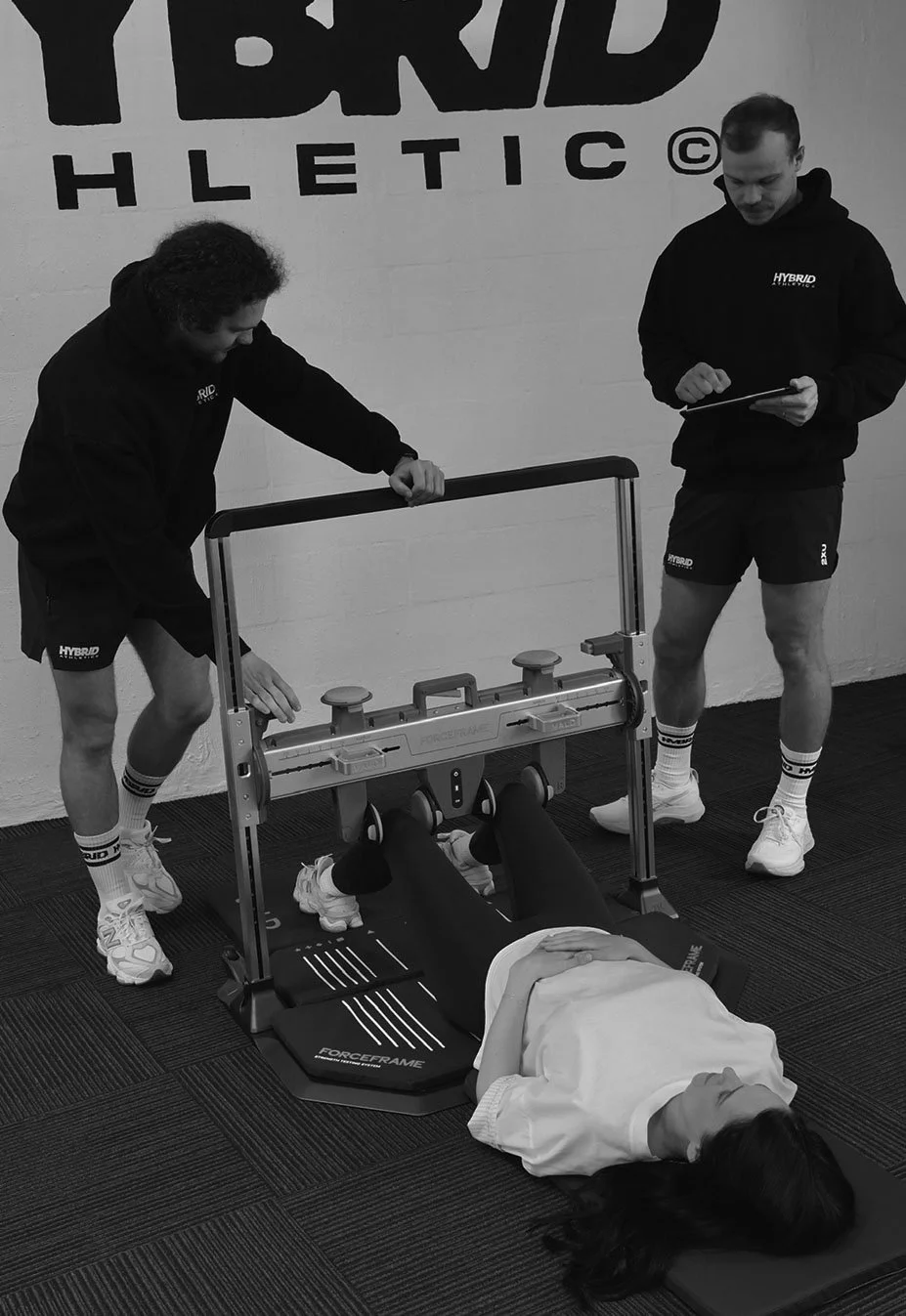 Two trainers working with a patient on a specialized rehabilitation machine in a gym. The patient is lying on her back on a mat, with her legs in the machine. The background features a wall with the words 'HYBRID HLETIC'.