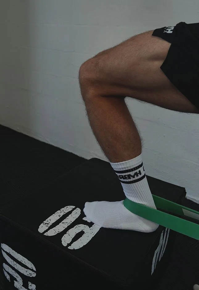 Close-up of a person's leg doing a physical exercise with a resistance band, on a black box with white text, against a white wall.