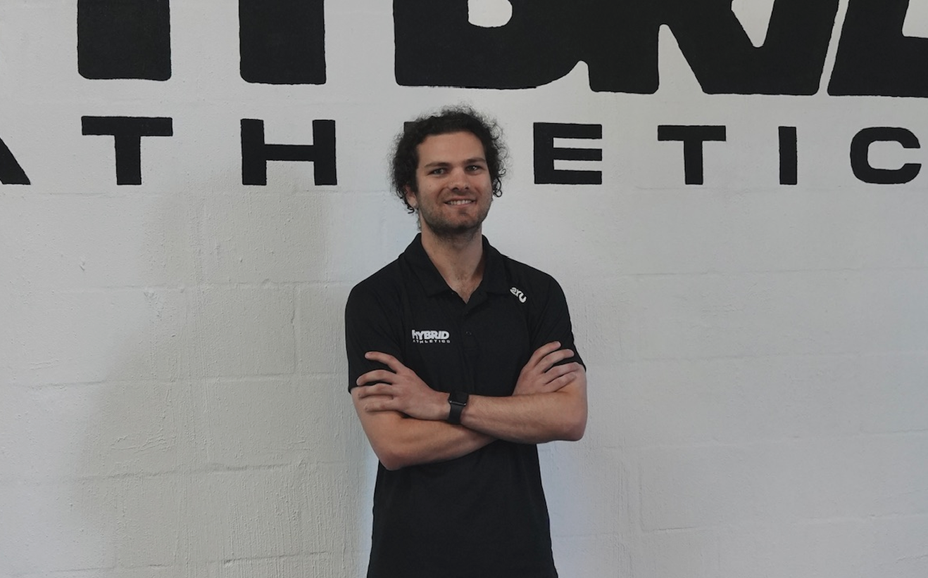 A man with curly hair and a beard smiling, standing with arms crossed in front of a white wall with black text.