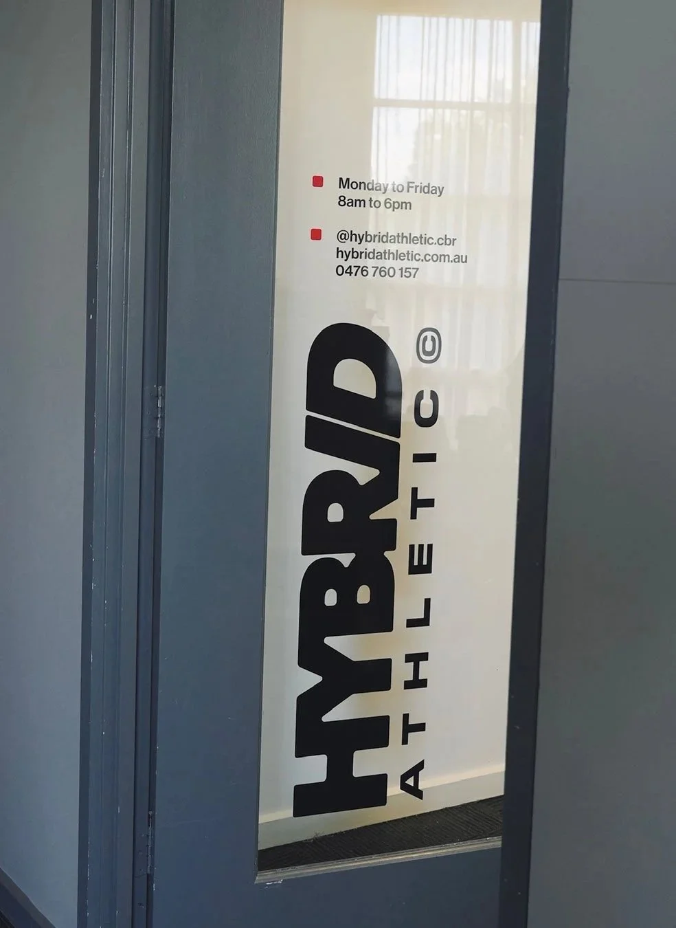Sign on a door for Hybrid Athletic, a fitness center, with the name in large black letters and hours from Monday to Friday 8am to 6pm. Contact information including Instagram handle, website, and phone number listed in smaller text.