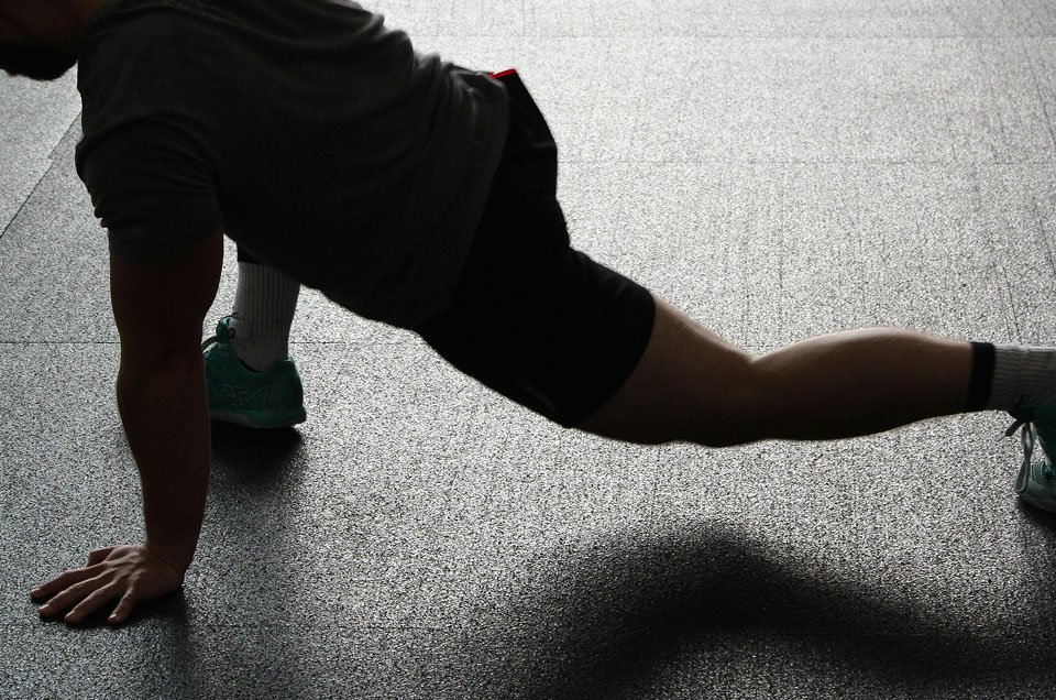 A person in athletic clothing performing a stretch on a gym floor, with one hand on the ground and one leg extended to the side.