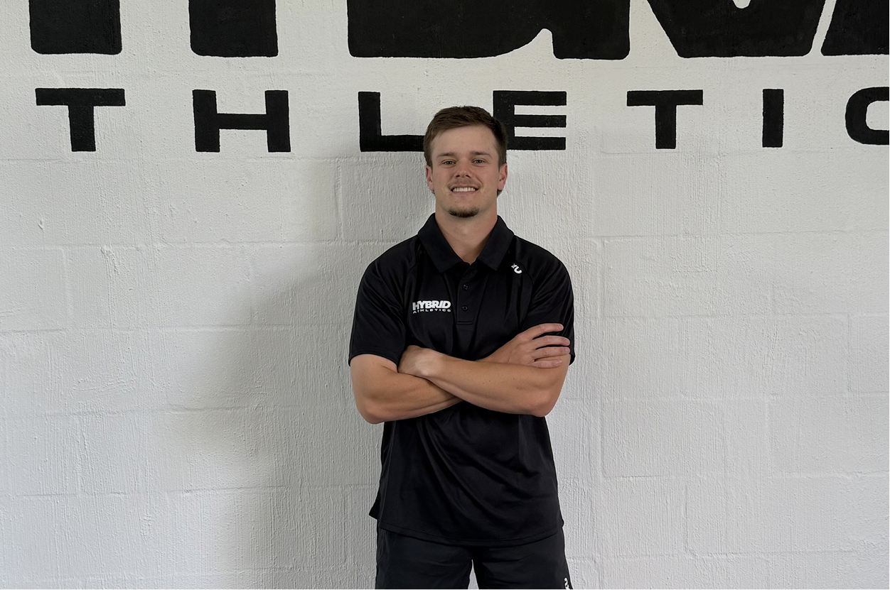 A young man with short brown hair, wearing a black Nike polo shirt with 'HYBRID ATHLETICS' embroidered on it, stands crossing his arms in front of a white wall with large black lettering. He is smiling.