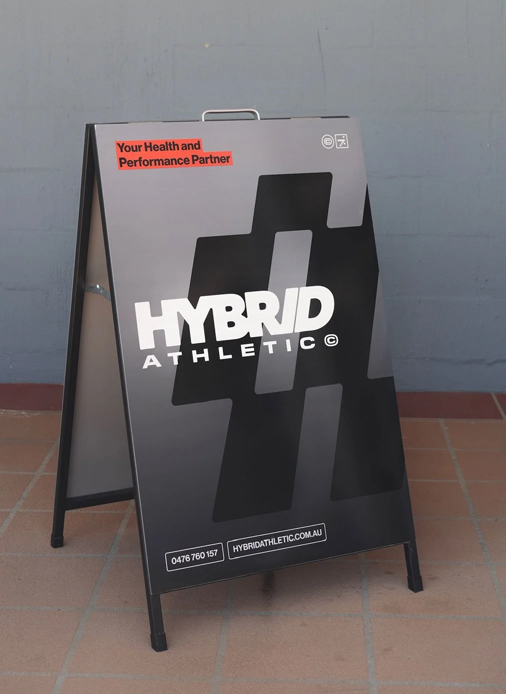 A black and gray A-frame sidewalk sign advertising Hybrid Athletic, with the text 'Your Health and Performance Partner' in red at the top, and a phone number and website at the bottom.