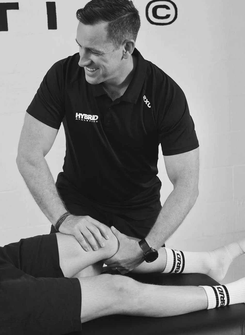 A male physiotherapist or trainer providing physical therapy or stretching treatment to a client's knee in a gym or rehabilitation setting. The professional is smiling and wearing a black polo shirt.