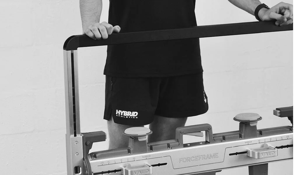 A person wearing black shorts and a black shirt standing behind a piece of fitness equipment, with their hands resting on the top bar.