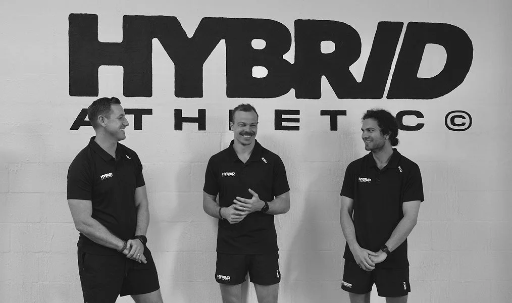 Three men standing in front of a wall with a large 'HYBRID ATHLETIC' logo, all wearing black shirts with matching logos, smiling and engaging with each other.