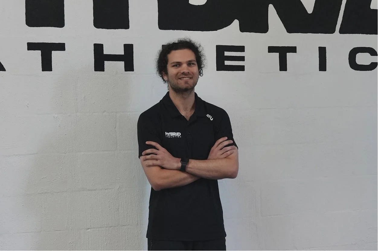 A man with curly hair and a beard standing with arms crossed, wearing a black polo shirt with logos, smiling, against a white wall with large black text.