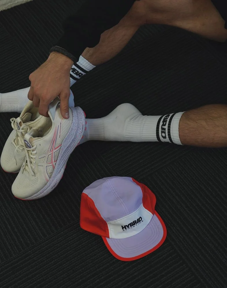 A person sitting on a dark carpeted floor, holding a pair of beige athletic shoes, with a white and red cap labeled 'HYBRID' nearby.