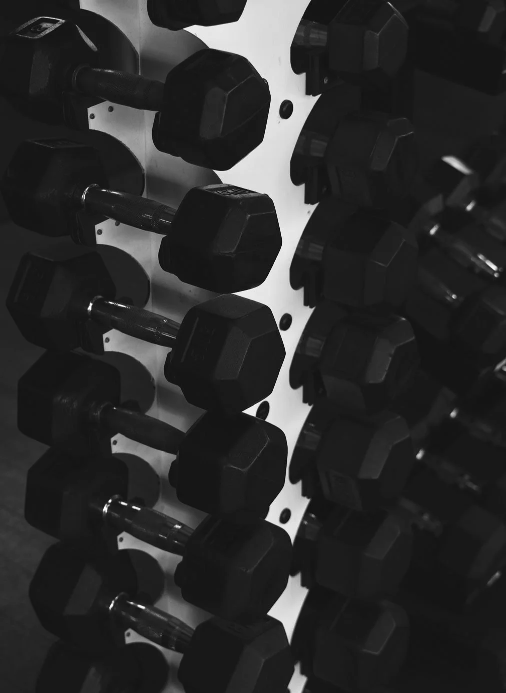 This is a black and white photograph of multiple dumbbells arranged on a rack, with the focus on the hexagonal-shaped weights.