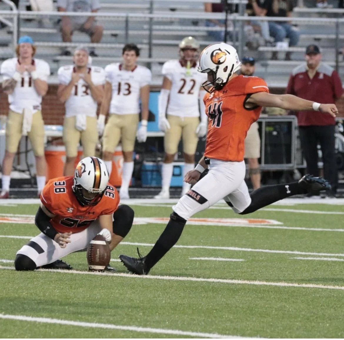 A football player in an orange jersey celebrating after a successful play, with one knee on the ground holding the football and another player running next to him; teammates in white and gold uniforms are in the background watching.