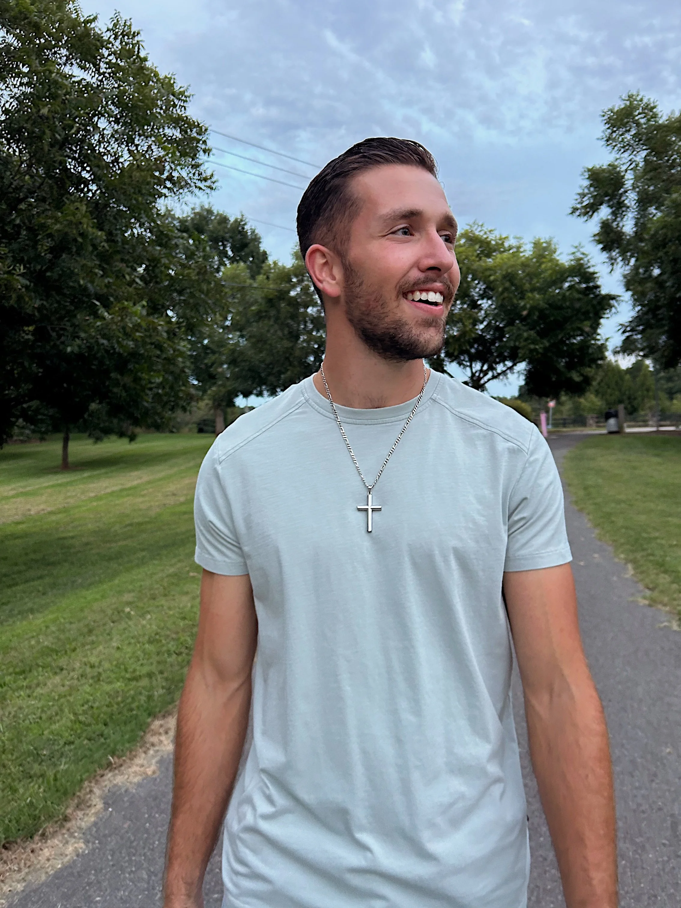 A smiling man with short dark hair and a beard wearing a light gray T-shirt and a necklace with a cross, standing outdoors on a paved pathway surrounded by green trees and grass under a partly cloudy sky.