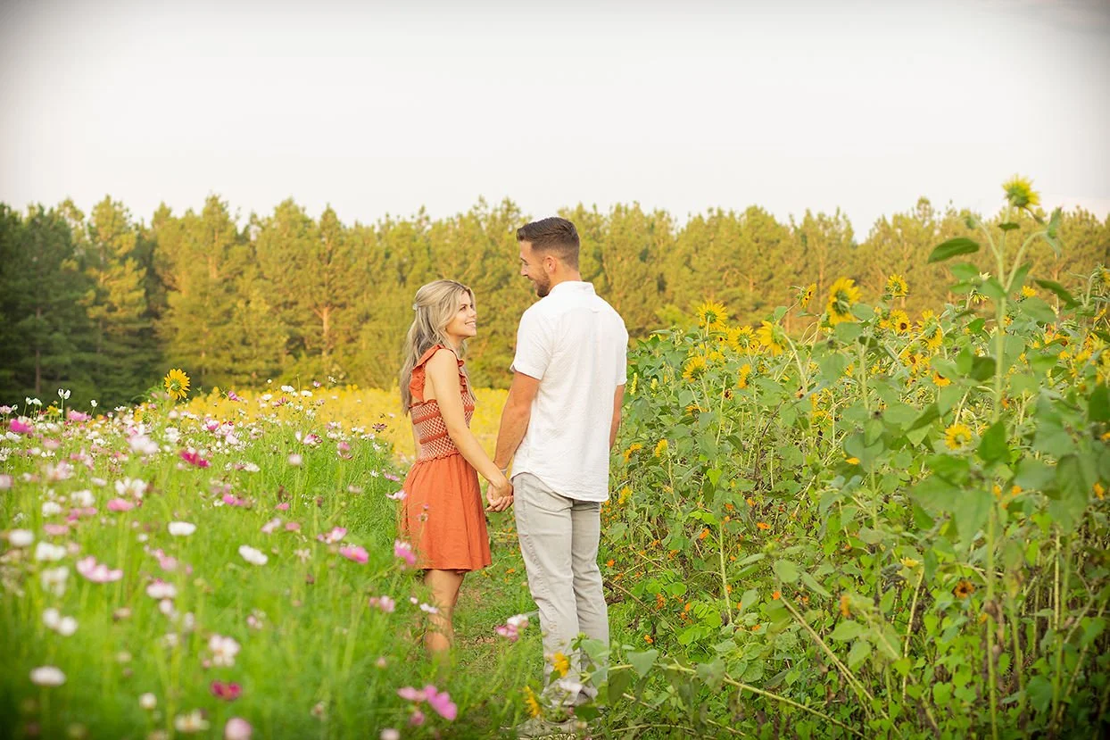 A couple stands hand in hand in a sunflower and wildflower field on a clear day, looking at each other and smiling, with trees in the background.