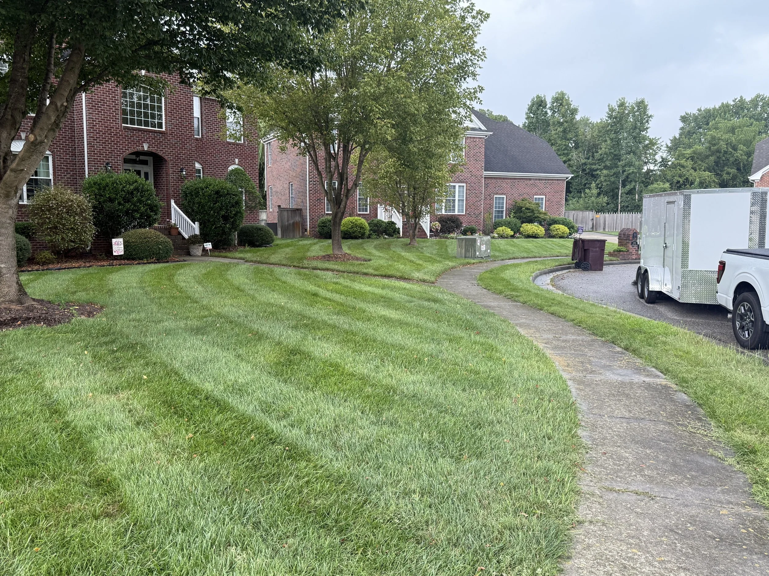 A residential sidewalk with well-maintained lawn and trees, leading to brick houses and a parking area with a white utility trailer and a pickup truck.