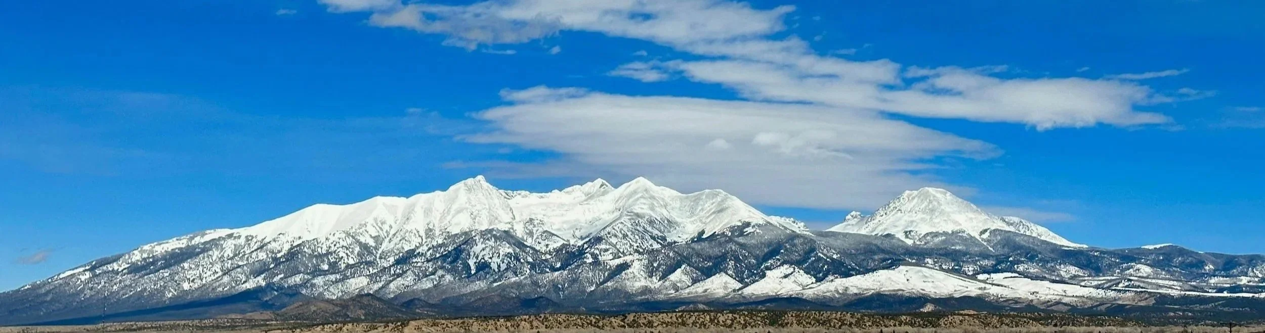 Snow-capped mountains under a partly cloudy blue sky with some green hills and desert terrain in the foreground.