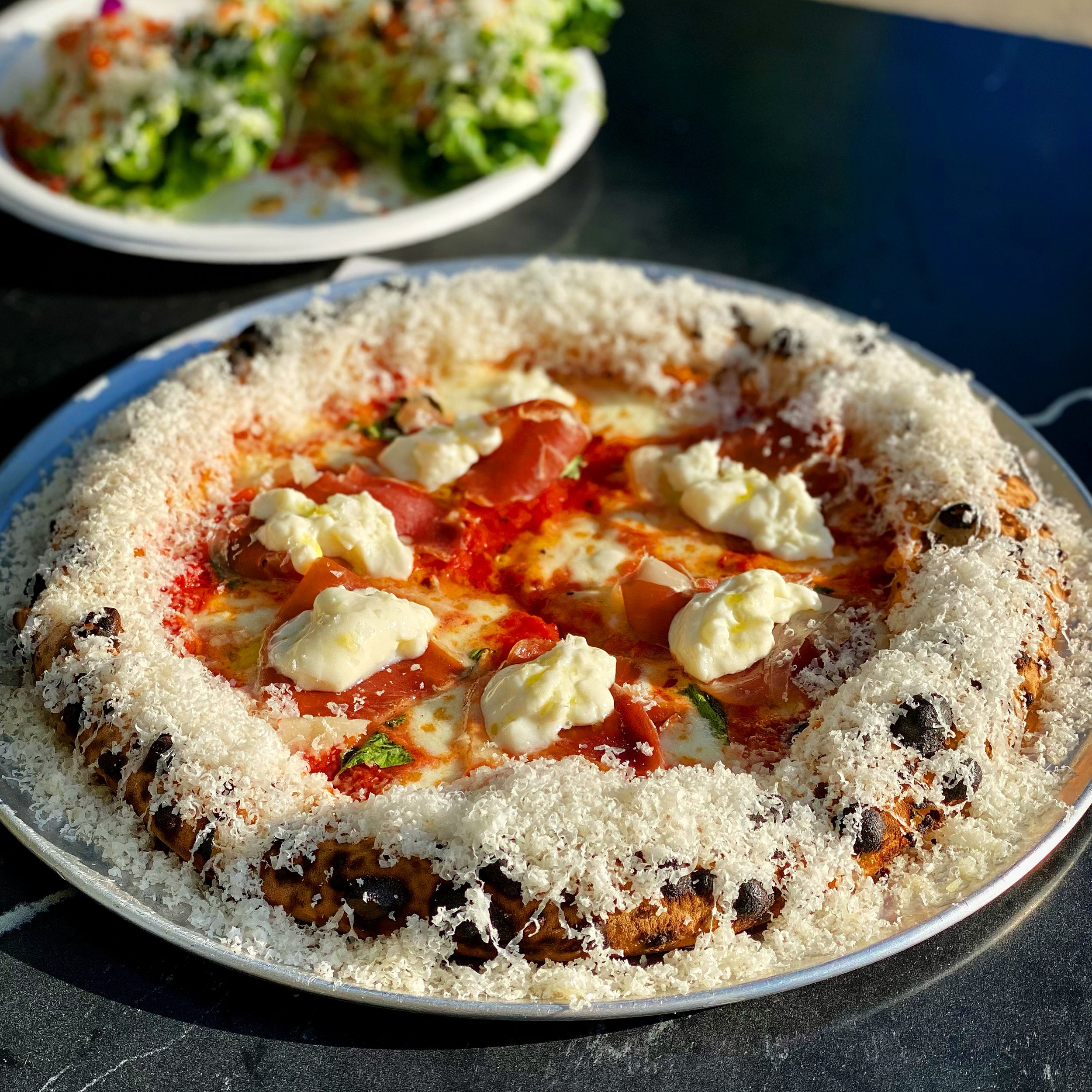 A pizza with a crust coated in grated cheese, topped with red sauce, cheese, sliced tomatoes, and green herbs, on a metal tray. In the background, a bowl of salad with vegetables.