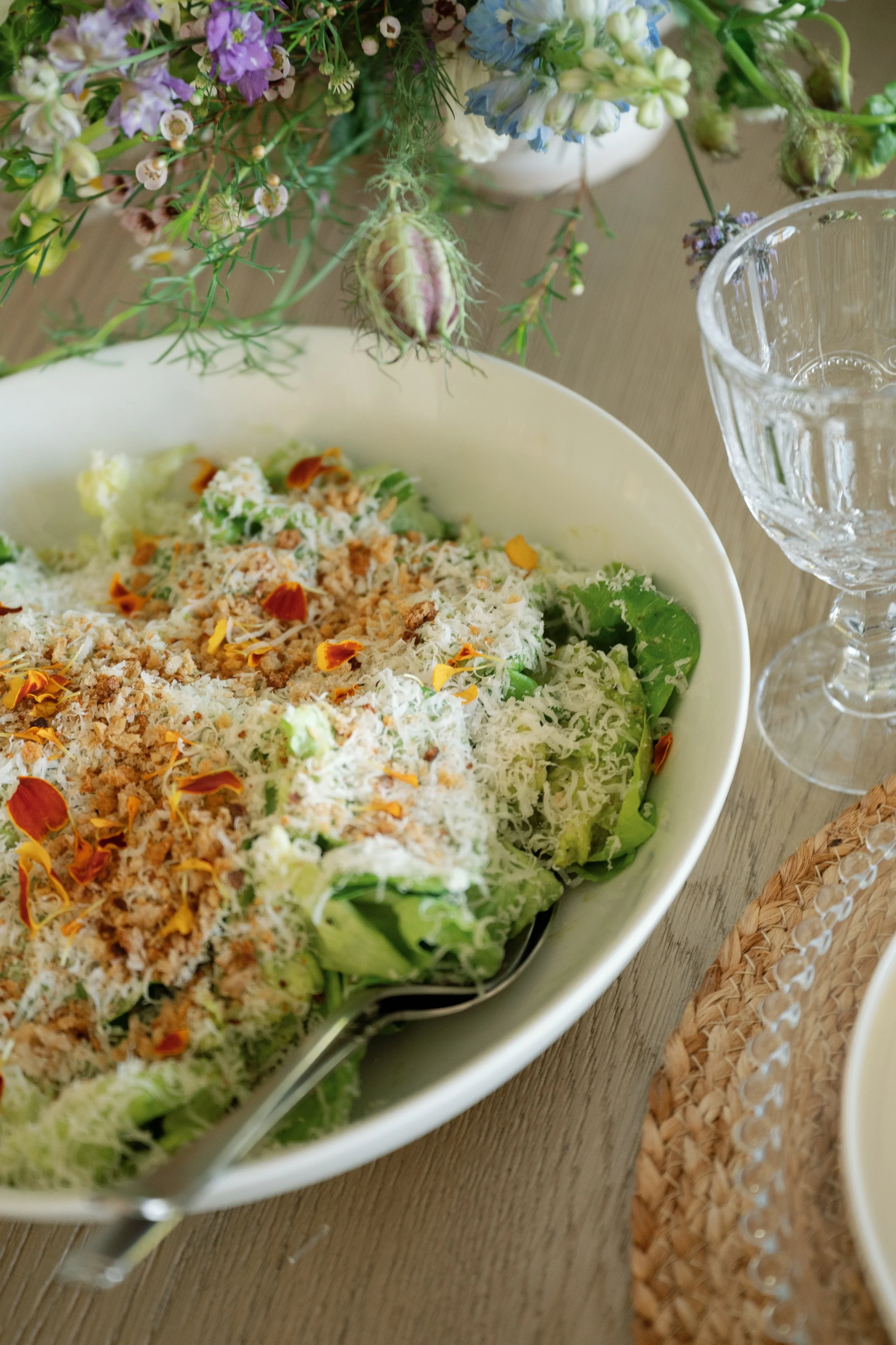 A salad with shredded cheese, chopped nuts, and edible flower petals in a white bowl, on a wooden table, with a floral arrangement and a clear glass pitcher nearby.
