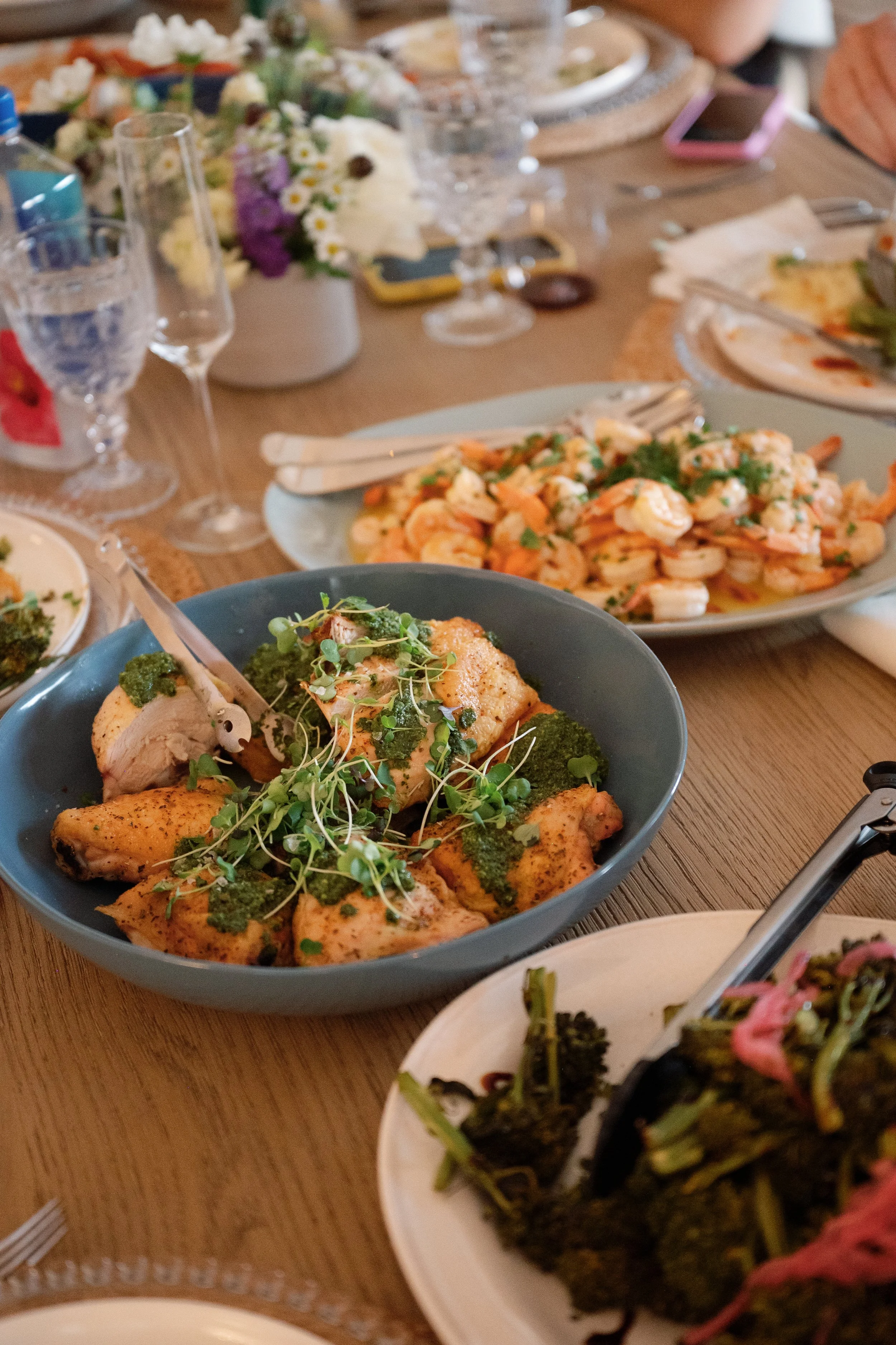 A table filled with various plates of food, including shrimp, grilled chicken with herbs and broccoli, and other dishes, along with glasses of water and a floral centerpiece.
