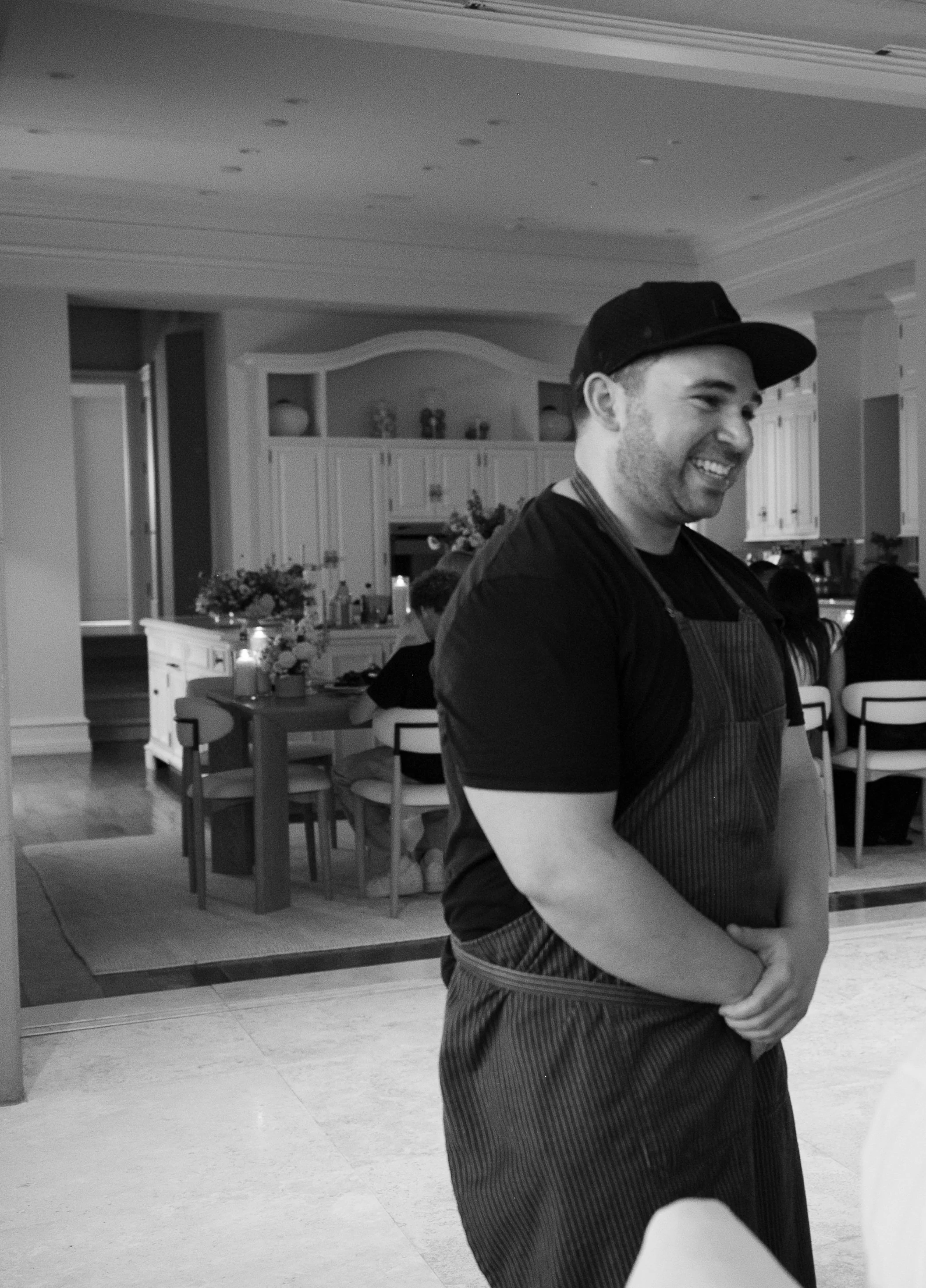 A man with a beard wearing a cap and an apron, smiling, standing indoors in front of a kitchen with white cabinets and a table with flowers, other people seated at the table.