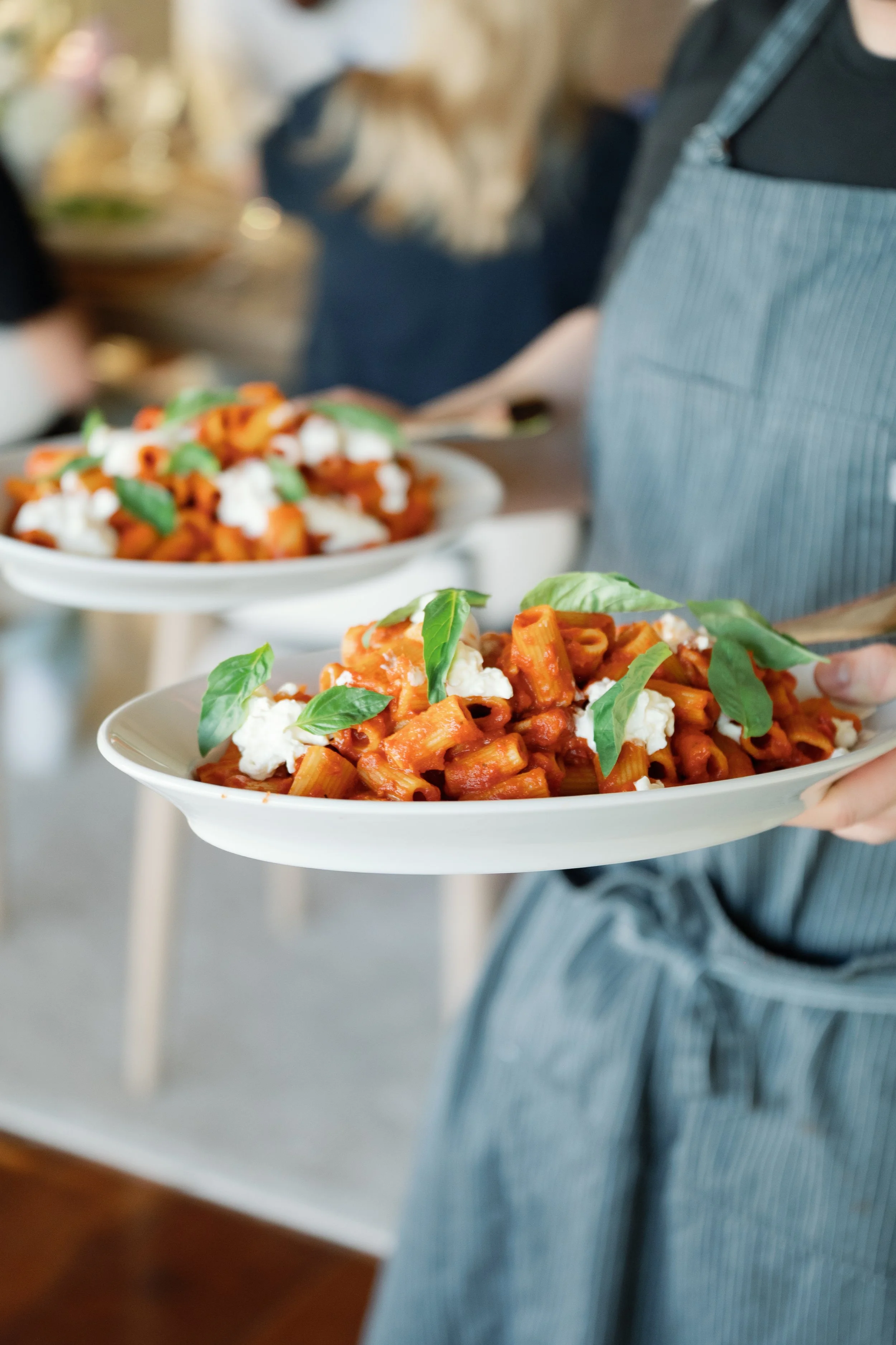 Person holding a plate of pasta with basil and cheese, several similar plates in the background.