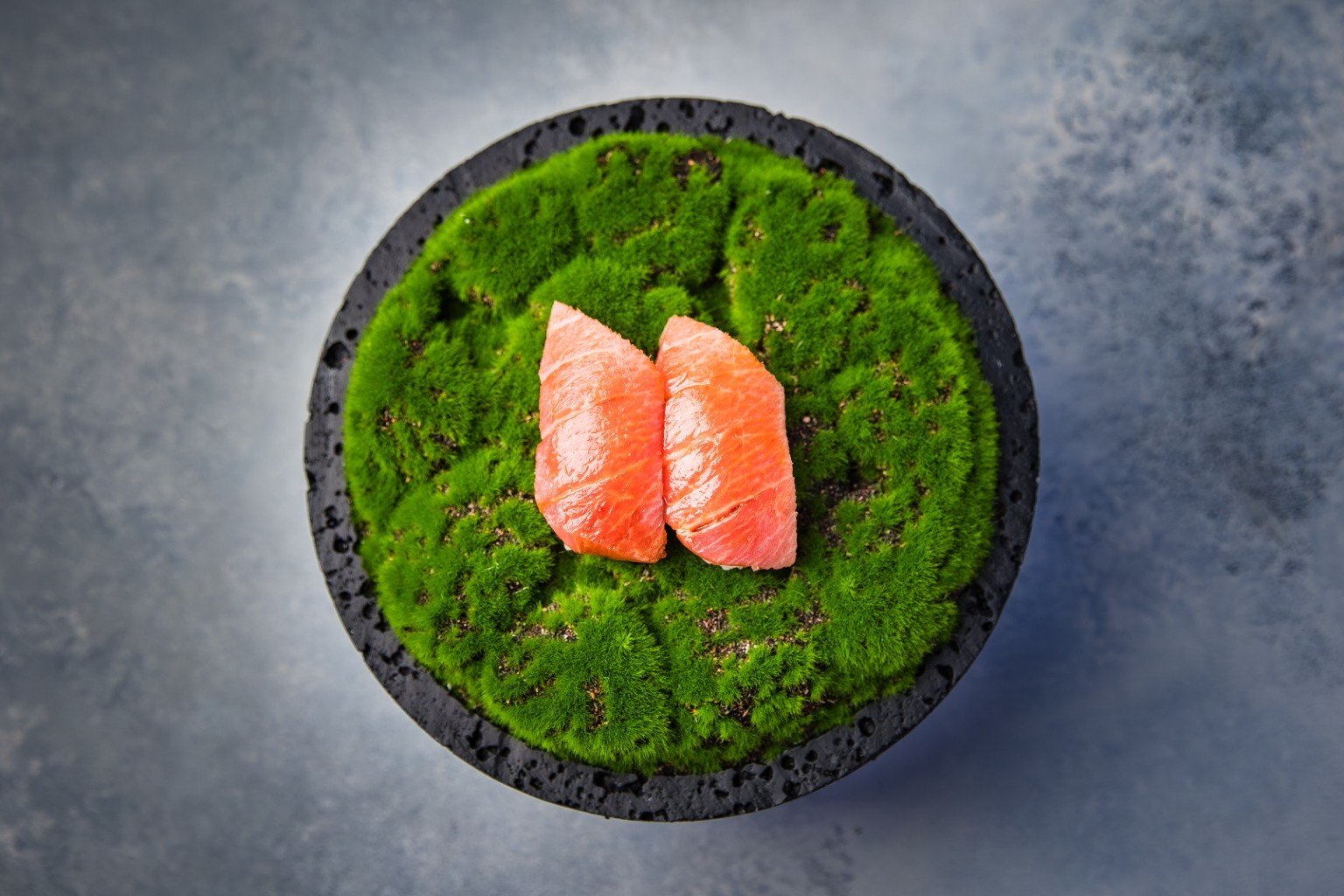 Two pieces of salmon nigiri presented on a moss-covered black plate by Chef Marco