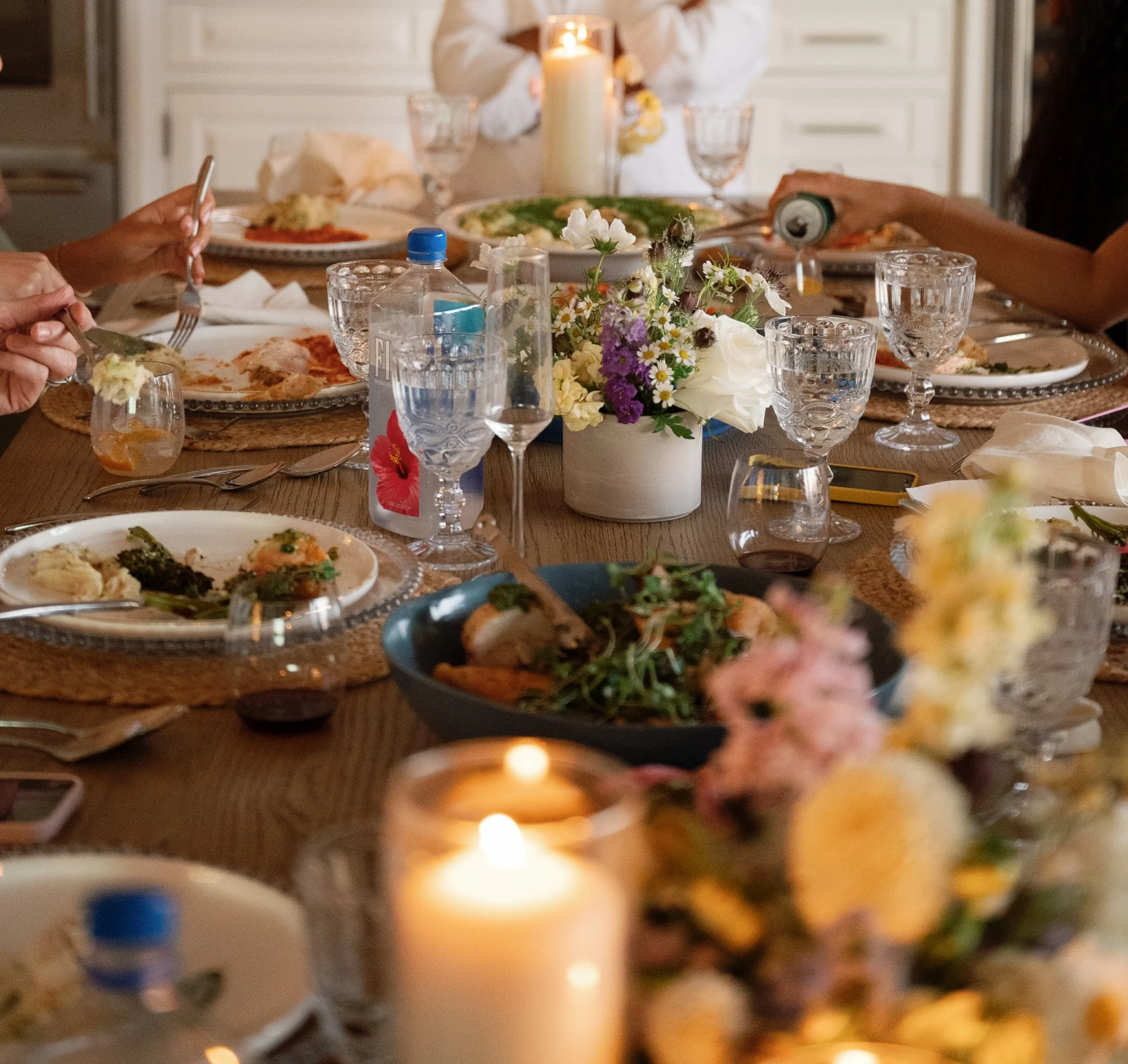 A dining table set for a meal with floral centerpiece, plates with food, water glasses, and napkins. In the background, someone is holding a drink can. The scene is illuminated by a candle on the table.