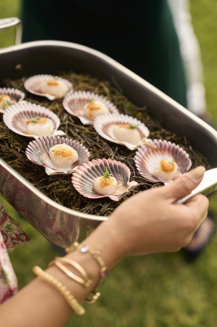 A person holding a silver tray with scallops in shells, garnished with herbs and sauce, on a bed of moss or seaweed outdoors.