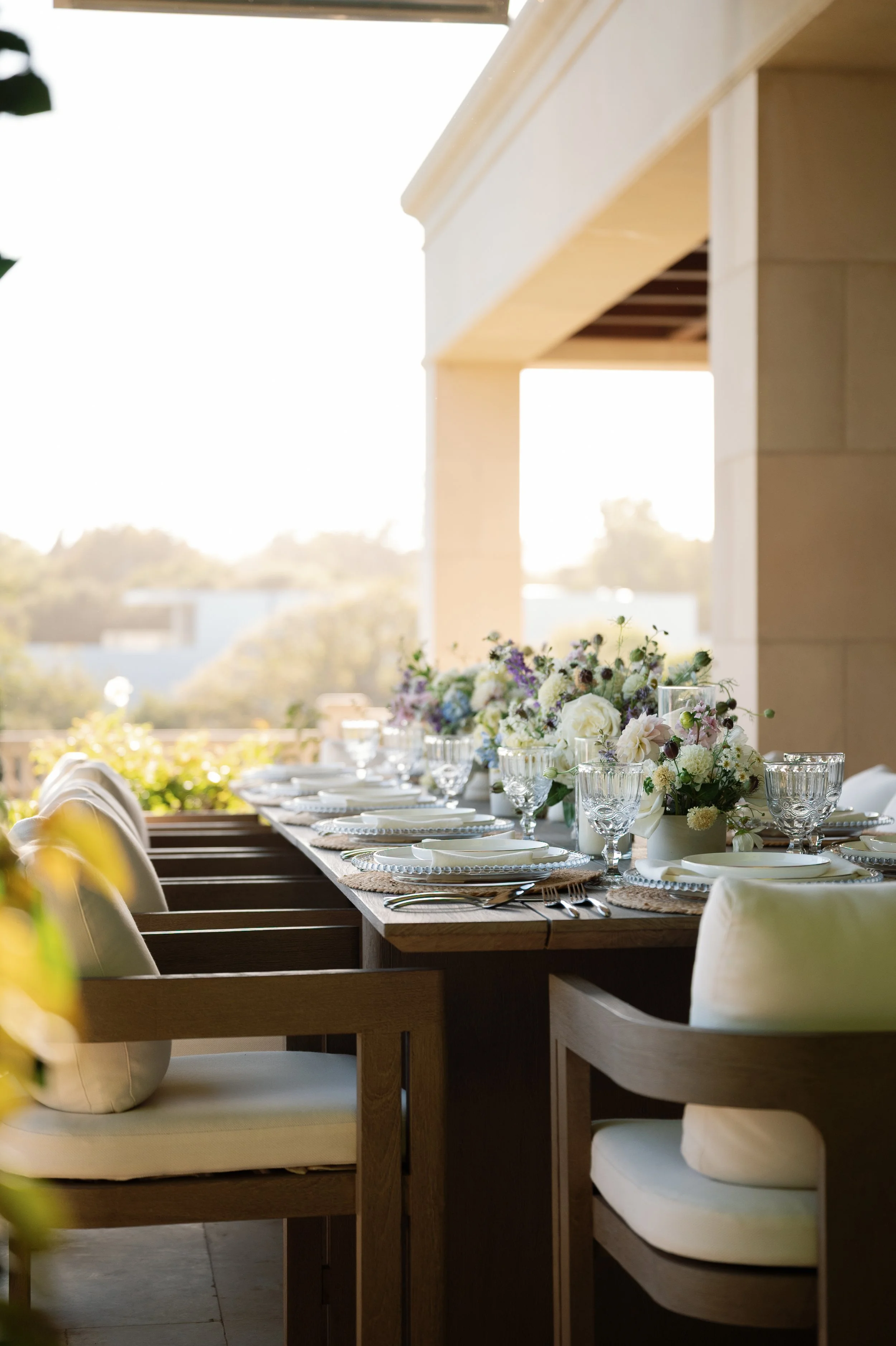 A beautifully set outdoor dining table with flowers and glassware, under a covered patio with a bright sky in the background.