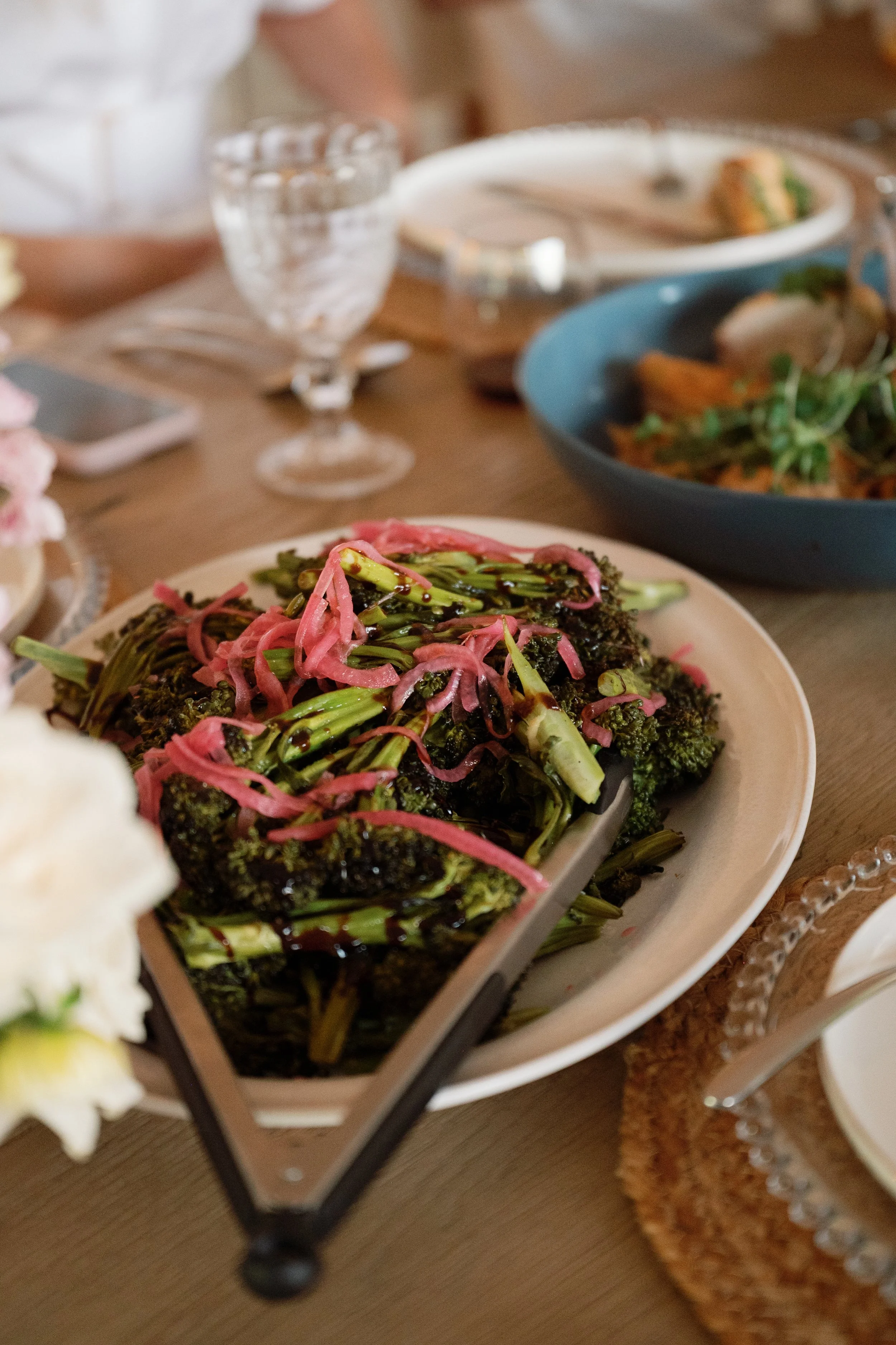 Close-up of a serving dish with roasted broccoli topped with pink pickled onion and sauce, on a table set for a meal with glasses, plates, and other food dishes in the background.